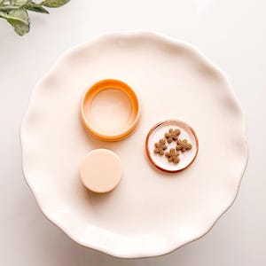 May include: A light peach-colored circular container with a matching lid and a small round plate containing four tiny brown gingerbread man-shaped cookies. The items are arranged on a large, scalloped-edge white plate with a smooth finish.