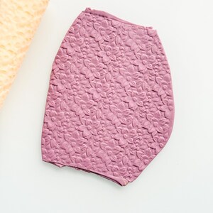 May include: A mauve-colored, floral-patterned object with a textured surface. The object is irregularly shaped and appears to be made of a pliable material. A cylindrical, light-yellow object is partially visible in the upper left corner.
