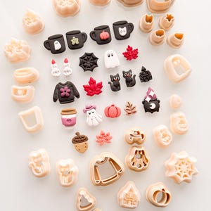 May include: A collection of beige plastic cookie cutters and small, colourful clay charms featuring Halloween and autumn themes. The charms include black mugs, ghosts, pumpkins, cats, sweaters, leaves, spiders, and a cauldron, arranged with matching cutters.