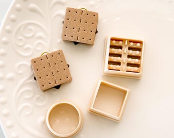 Könnte beinhalten: Draufsicht auf Miniatur-S'mores-Anhänger und -Behälter. Die Anhänger zeigen beige Graham Cracker mit dunkler Schokolade und Marshmallow-Füllungen. Die Behälter sind beige und quadratisch, einer offen und einer mit einem Gittermuster.