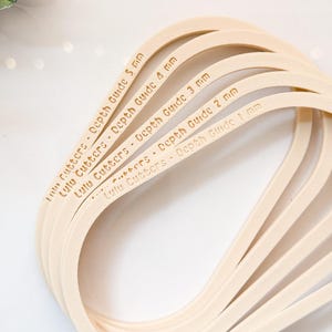 May include: A collection of beige Lulu Cutters depth guides, measuring from 1 mm to 5 mm. Each guide is marked with its depth in millimetres. The guides are curved and arranged in a flowing, overlapping design.