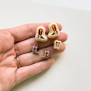 May include: A collection of tan and pink cowboy boot-shaped cookie cutters. The cutters are in various sizes, with some featuring pink bows. The boots have detailed cut-out designs. The items are displayed on a light background.
