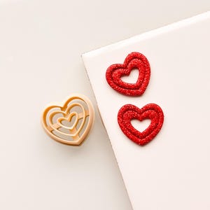 Puede incluir: Dos formas de corazón de purpurina roja y un cortador de galletas en forma de corazón de plástico beige sobre una superficie blanca.