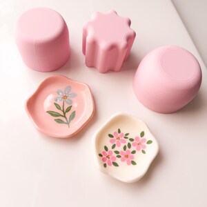 May include: Three pink containers and two small dishes. One container is cylindrical, one is fluted, and one is rounded. The pink dish features a single blue flower, while the cream-coloured dish displays a cluster of small pink flowers with green leaves.