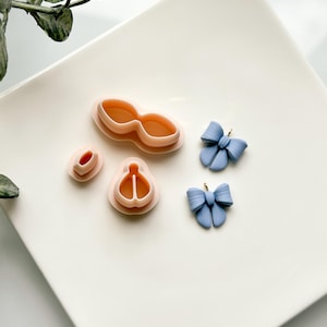 May include: A set of three pink plastic cookie cutters shaped like a bow, a heart, and a pair of lips. Two blue polymer clay bows with gold jump rings are also shown.