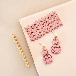 Puede incluir: Pendientes de arcilla con estampado rosa y morado con detalles dorados. Los pendientes están hechos de dos cuadrados de diferentes tamaños. Los pendientes están sobre una superficie blanca.