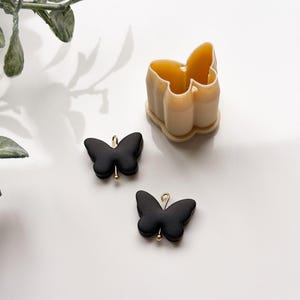 May include: A butterfly-shaped cookie cutter and two black butterfly charms with gold-colored findings. The cookie cutter is light beige and has a butterfly shape. The charms are matte black.