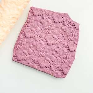 May include: A close-up shot of a textured, mauve-colored floral pattern. The design features raised flowers and leaves. A piece of light beige, textured material is in the upper left corner. The background is white.