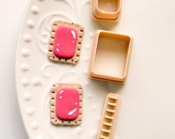 Könnte beinhalten: Zwei Miniatur-Kekse mit rosa Zuckerguss und weißen Akzenten, zusammen mit drei beigefarbenen Ausstechern. Die Artikel sind auf einem weißen Teller mit dekorativem Muster angeordnet. Die Ausstecher sind rechteckig und haben ein glattes Design.
