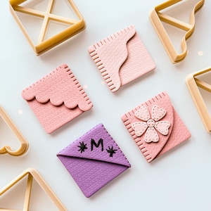 May include: Overhead view of craft supplies: light brown plastic geometric shapes and pink and purple decorative squares. The squares feature stitched edges, scalloped edges, and a flower design. One square is purple with the letter "M" and asterisks.