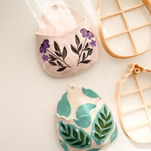 May include: Two ceramic wall pockets and two plastic cookie cutters. One wall pocket is light pink with purple flowers and black stems. The other is cream with teal shapes, green leaves, and white dots. The cutters are tan and egg-shaped.
