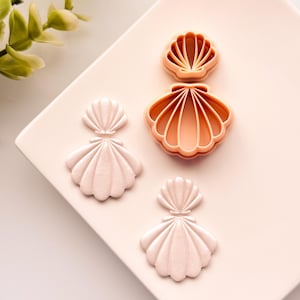 May include: A collection of seashell-shaped cookie cutters and finished earrings. The cutters are peach-colored, while the earrings are a pale pink. The items are arranged on a light pink square plate, with a sprig of green leaves in the background.