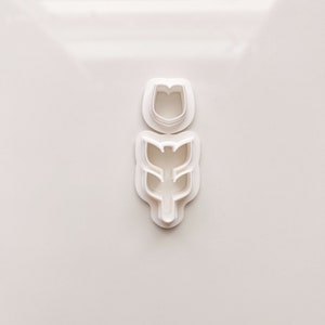 May include: Two white plastic flower-shaped cookie cutters. One cutter is a simple tulip shape, and the other is a tulip with a stem and leaves. The cutters are on a white background.