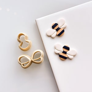 May include: A set of three cookie cutters, two are shaped like bees and one is shaped like a bow. The bee cookie cutters are white with black stripes and a yellow body. The bow cookie cutter is a simple shape.
