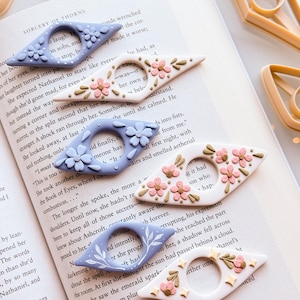 May include: Handmade book page holders in shades of white and lilac, adorned with floral and leaf patterns. These decorative reading accessories are ideal for marking pages in books.