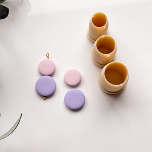 Puede incluir: Vista superior de artículos de artesanía. Se ven tres cortadores cilíndricos beige y varias piezas redondas de arcilla en colores pastel, lavanda y rosa. Una pieza de arcilla tiene una cuenta dorada adjunta. Los artículos están dispuestos sobre una superficie blanca.