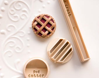 Puede incluir: Una tarta en miniatura con una corteza de rejilla y un juego de herramientas para hacer pasteles de color beige. Las herramientas incluyen un cortador de corteza de pastel, un sello redondo con líneas paralelas y un cortador redondo con las palabras "pie cutter".