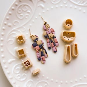 Puede incluir: Un par de pendientes de arcilla polimérica patchwork hechos a mano con postes dorados, con cuadrados rosas, morados y azul marino. También se ven varios cortadores de arcilla beige de varias formas.