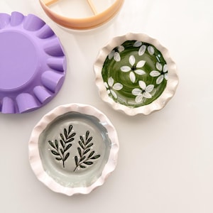 May include: Two small, round, scalloped-edge ceramic dishes featuring hand-painted designs: one with white flowers on a green background and one with dark green leafy sprigs on a light gray background. Includes a purple mold and a peach-colored divider.