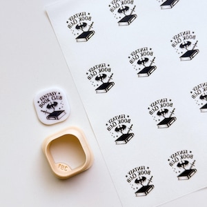 May include: A sheet of white stickers with a black graphic of a book, bat, and sword, with the text "FANTASY BOOK CLUB". A square cookie cutter and a single sticker are also present. The cookie cutter has the letters "FBC" on it.