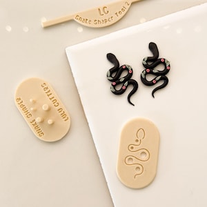 May include: A collection of polymer clay tools and snake-themed earrings. The tools include a beige snake shaper, a cutter with the text "Lulu Cutters Snake Shaper", and a stamp. Two black snake earrings with pink and green floral details are also present.