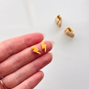 May include: Two small yellow lightning bolt shaped earrings. The earrings are held in a person's hand.
