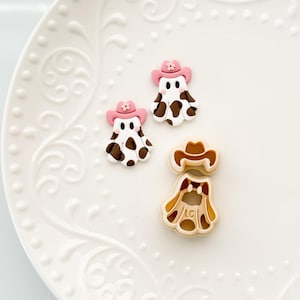 Puede incluir: Tres elementos decorativos: dos figuras con forma de fantasma con manchas marrones y blancas, que llevan sombreros de vaquero rosas con detalles florales, y un cortador de galletas con forma de vaquero. Los artículos están sobre un plato blanco estampado.