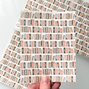 May include: A sheet of stickers featuring a repeating pattern of illustrated bookshelves. The books are in shades of brown, beige, and gray, with some featuring leaf designs. The stickers are held by a hand, with additional sheets visible in the background.