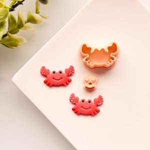 May include: Two red plastic crab cookie cutters with a white background. The cutters are in the shape of a crab with a smiling face and white dots. There is also a larger, single-piece crab cutter in the background.