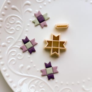 May include: A white ceramic plate with embossed floral patterns displays three small, colourful star-shaped decorations and two cookie cutters. The decorations are purple, green, and pink, resembling quilt blocks. The cutters are beige.