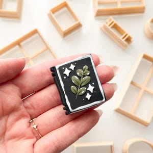 May include: A small, rectangular black book-shaped charm featuring a central green leafy branch illustration, white star accents, and a white border. The charm is surrounded by several light tan, geometric plastic cookie cutters.