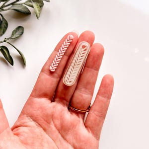 Puede incluir: Dos piezas decorativas de arcilla polimérica con forma de trenza, una en una combinación de colores rosa claro y blanco, y la otra transparente. Las piezas se sostienen en una mano, con un anillo de oro en el dedo anular. Una ramita de vegetación está en el fondo.