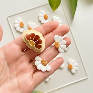 Puede incluir: Un conjunto de pendientes de arcilla polimérica en forma de margarita blanca con un centro amarillo. Los pendientes se muestran junto a un cortador de galletas en forma de margarita.