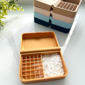 Puede incluir: Un recipiente de plástico amarillo con una tapa con bisagras. El recipiente tiene dos compartimentos, uno con un patrón de cuadrícula y el otro lleno de polvo blanco.