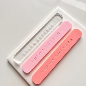 Puede incluir: Dos rodillos de acrílico con el texto "Lulu & Cutters" en relieve. El rodillo superior es transparente y el rodillo inferior es rosa.