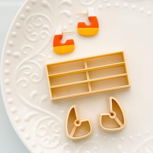 Puede incluir: Pendientes en forma de caramelos de maíz con postes dorados, junto con un molde rectangular y dos cortadores de caramelos de maíz. Los pendientes presentan secciones naranja, amarilla y blanca. Los artículos están dispuestos sobre un plato blanco.