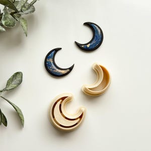 May include: Two crescent moon cookie cutters, one in a light beige color and the other in a dark blue color with gold accents. The blue moon cutter is on a white surface.