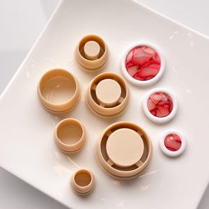 May include: A set of six round, beige plastic molds for making resin crafts. The molds are of varying sizes and are arranged on a white plate. There are also three small, round, red and white resin cabochons with gold flecks.
