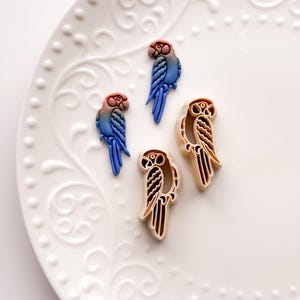 May include: Four parrot-shaped cookie cutters. Two are blue, red, and pink, and two are tan with brown details. The cutters are arranged on a white, patterned plate, ideal for baking themed cookies.