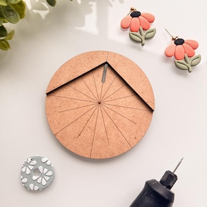 May include: A collection of craft supplies and handmade jewelry. Includes a wooden circle with radial lines, a gray and white floral earring, a pair of flower earrings with peach petals, and a black and gray rotary tool.