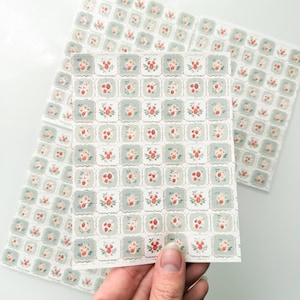 May include: A sheet of square stickers featuring a repeating pattern of small red strawberries with green leaves on a light sage green background. The stickers have scalloped edges and are arranged in a grid format on a white backing sheet.