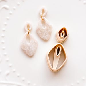 May include: Two pairs of earrings and two clay cutters are displayed on a white plate. The earrings have a marbled, off-white appearance with gold-colored findings. The clay cutters are beige and diamond-shaped, with a central oval cutout.