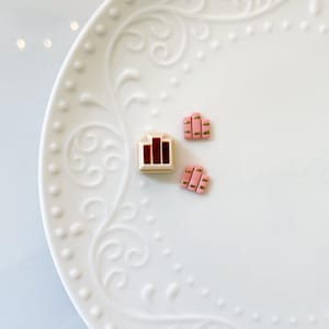 May include: Three small, decorative items on a white, patterned plate. One is a beige rectangle with three vertical red lines. The other two are pink rectangles with gold accents, resembling miniature stacks.