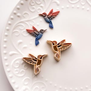 May include: Four hummingbird-shaped decorations. Three are colorful with red, blue, and brown hues, while the other two are brown and beige. They are arranged on a white, patterned plate.