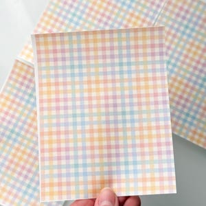 May include: Square paper sheets with a pastel plaid pattern in shades of pink, yellow, blue, and orange. The sheets are held by a hand, with others visible in the background. The design is a repeating gingham check.