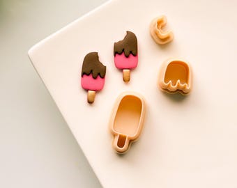 May include: Two pink and brown ice cream pops with a bite taken out of one, and three plastic cookie cutters in the shape of ice cream pops on a white surface.