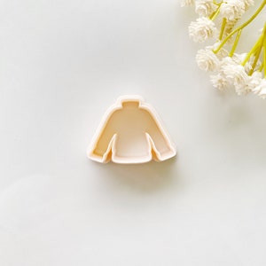 Puede incluir: Un cortador de galletas de plástico con forma de suéter. El cortador es de color melocotón claro.