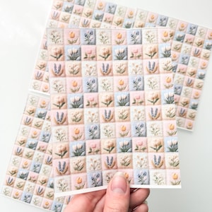 May include: A rectangular card featuring a grid pattern of small squares, each containing an embroidered-style illustration of various flowers in soft pastel shades of pink, blue, yellow, and white against a light background.