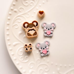 Puede incluir: Una colección de cortadores de arcilla polimérica marrón claro y gris en varias formas, incluyendo una cara de oso, un corazón y dos ratones grises sosteniendo corazones rosas. Los cortadores están dispuestos sobre un plato blanco con dibujos.