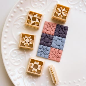 Könnte beinhalten: Eine Sammlung beigefarbener quadratischer Ausstecher mit verschiedenen geometrischen Designs, darunter Sterne und Dreiecke, neben einem bunten Patchwork aus Keksen in Rosa, Blau und Lila. Die Artikel sind auf einem weißen, dekorativen Teller angeordnet.